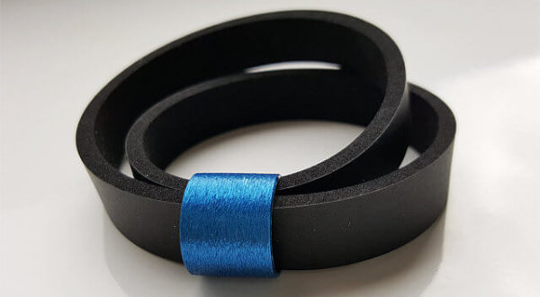 Doppelreihiges Neoprenarmband in einem Stück, wobei das Band zweimal durch ein blauglänzendes, metallenes, zylinderförmiges Schmuckaccessoire gezogen ist.